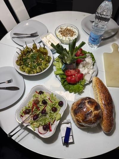 The image shows a table laden with food. There are two types of bread, one long and one round, both wrapped in plastic wrap. A plate holds cut tomatoes, sliced onions, lettuce, spring onions, and cucumber slices. Another dish contains a white creamy substance topped with nuts. A container of white cheese cubes is also visible. A plate is filled with green peppers, garnished with herbs. A salad with cabbage, red peppers, radishes, olives, and tongs is also present. There are plates with silverware set for dining. A bottle of water sits on a plate. A pack of cigarettes and a lighter are next to one of the breads. Given the typical food composition, this scene appears to be taking place in North Macedonia. Specific details, such as local food preparations, suggest it might be located in the city of Skopje. There are no people explicitly visible in the image. The setting suggests a casual meal or gathering.
