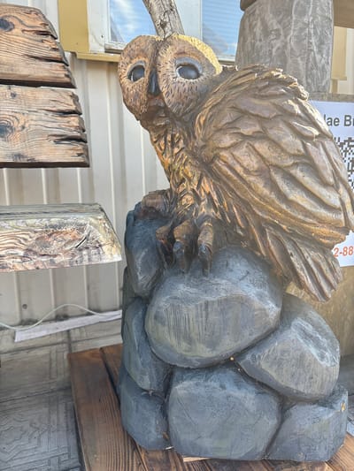 An outdoor scene in Vyra, Russia, features a detailed wooden sculpture of an owl perched on a cluster of rocks. The owl is carved from light brown wood with darker brown accents, and its feathers are intricately rendered. Its eyes are large and black, giving it a watchful expression. The rocks beneath it are carved from darker gray wood, with rough textures suggesting natural stone.

To the left of the sculpture, there are stacked wooden elements that appear to be part of a rustic structure or display. These are also made of aged, weathered wood with visible grain patterns and some signs of wear. Behind the sculpture, a light-colored corrugated metal wall is visible, and a window with a white frame is present in the upper section of the wall. A QR code and some text are also visible on a white sign to the right of the owl sculpture. The ground beneath the sculpture is a wooden platform composed of several planks. The overall impression is that of a market stall or an outdoor craft display.