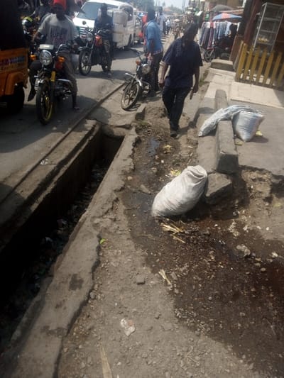 A street scene depicts a deep, open drainage ditch filled with trash beside a deteriorating sidewalk. The road is congested with motorcycles, cars, and pedestrians. Two large white bags sit on the sidewalk, partially blocking the pedestrian path. The scene suggests poverty and neglect, with visible disrepair in the infrastructure and a general lack of cleanliness. A man walks on the sidewalk near the bags.