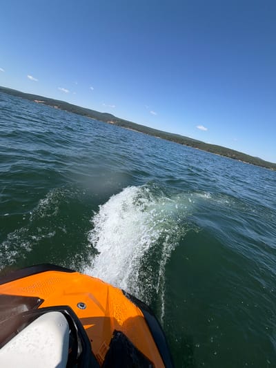 An eye-level, wide-angle shot captures the front portion of an orange jet ski on a body of water. The jet ski is moving, evidenced by the white spray and wake trailing behind it. The water is a deep green, with visible ripples and small waves. In the distance, a tree-covered shoreline curves around the water under a clear blue sky with a few scattered white clouds. The image appears to be taken in Dolni Pasarel, Bulgaria, on a bright, sunny day.