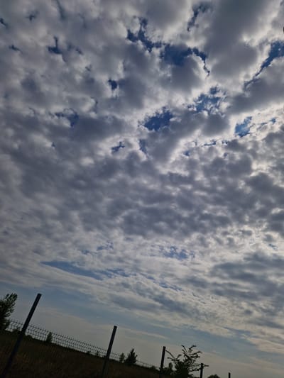 The image depicts an outdoor scene with a fence in the foreground and a cloudy sky filling the majority of the frame. The fence is made of dark metal posts and mesh, with a few scattered trees visible behind it. The sky is filled with a mix of dark and light clouds, with patches of blue sky visible through the gaps. The overall impression is one of a cloudy day with a rural or semi-rural setting, possibly on the outskirts of the city of Bahovitsa, Bulgaria. No people or specific actions are visible in the image.