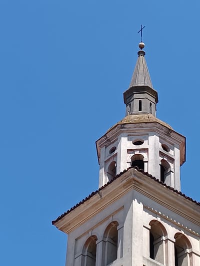 The image shows a portion of the top of the bell tower of the church of Mezzana Sant'Antonio, Italy, set against a clear blue sky. The tower is primarily white with a grey spire topped with a cross. The structure consists of multiple levels with arched openings and decorative elements. The bottom portion of the tower visible features arched windows, while the upper levels have smaller, round or rectangular openings. The building is old, with some signs of weathering.