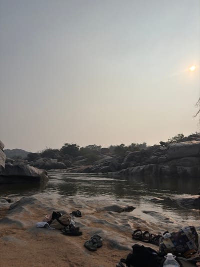 The scene depicts a rocky river environment in Hale Bandi Harlapura, India, under a hazy, light grey-beige sky with a prominent sun in the upper right quadrant. The foreground features a sandy shore with light-colored, undulating rock formations bordering a body of water. On the left side of the foreground, a light-colored garment, a tan fabric item, one pair of black sandals, and one pair of olive green sandals are placed on the sand. On the right side of the foreground, a patterned fabric bag, a dark fabric item, a clear plastic water bottle with a white cap, and one pair of dark sandals are present. The extreme right edge of the frame shows dark, curly hair, indicating the presence of an individual partially outside the frame. The midground is occupied by a calm river with a dark, reflective surface, flowing between prominent rock formations. The background is dominated by numerous large, grey boulders and rock outcrops extending into the distance on both sides of the river, interspersed with sparse green vegetation.