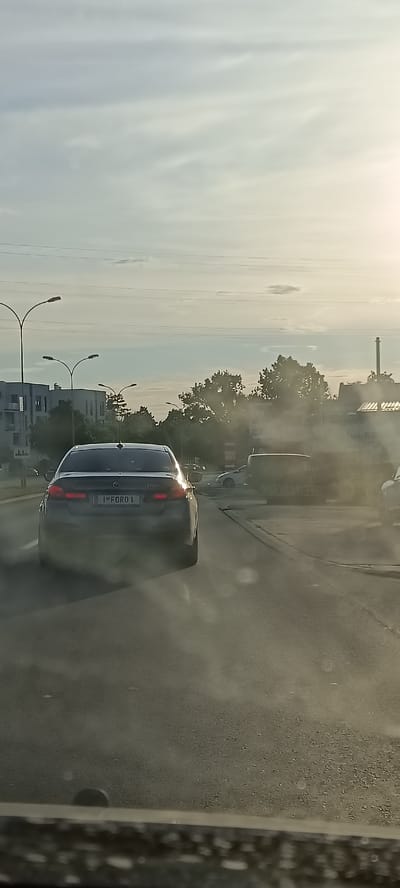 The image shows a rear view of a dark-colored sedan driving on a road in Veternik, Serbia. The car has a license plate that reads "1 FORO 1". The sky is overcast with some sunlight breaking through. There are trees and buildings visible in the background, as well as streetlights. The road surface appears to be asphalt. There is some light haze or dust in the air, particularly around the car. The image appears to be taken from another vehicle.