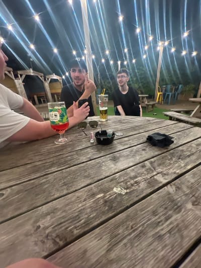 Three people sit at a wooden table under string lights. A glass of beverage with tropical print, another taller glass with beer, sunglasses, an ashtray and a pair of black gloves occupy the table. One person in a black shirt is taking a picture with their phone. Another person in black stares towards the camera. One person is wearing a white t-shirt.