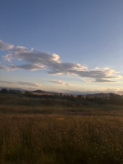 The image depicts a serene landscape under a partly cloudy blue sky, possibly taken at dusk or dawn. The foreground is dominated by a field of tall, dry grass, illuminated by soft, warm light, suggesting it's either late afternoon or early morning. In the middle ground, there's a line of dark green trees and shrubs, behind which lie fields of golden-brown crops and gentle rolling hills. Further in the distance, a series of mountains form the horizon, their peaks softened by the atmospheric light. Several clouds are scattered across the sky, some appearing white and fluffy, while others are tinged with the warm colors of the setting or rising sun. A single, small dark object, potentially a bird or a distant aircraft, is visible in the sky above the right side of the hills. The overall impression is one of natural beauty and tranquility. The image is located in the vicinity of Naiden Gerovo, Bulgaria.