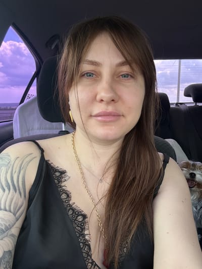 A selfie of a woman with brown hair and green eyes sitting in a car. She is wearing a black top with lace trim and a gold necklace. A small dog is visible behind her head. The sky outside the car window is light purple. The location is potentially Grushevskaya, Russia, though no visual elements confirm this.