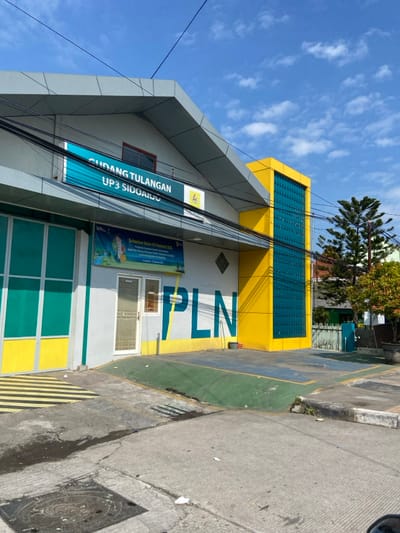 The image shows a building located in the city of Kecamatan Tulangan, Indonesia, with coordinates -7.481, 112.645. The building is a single-story, and is painted with yellow and turquoise. It has a sign that reads "Gudang Tulangan UP3 Sidoarjo". There is a large, white "PLN" painted on the side of the building. The building has a large, open doorway leading to the interior. The door is a glass and metal door, framed in white, and has a few small, black trash cans and a planter in front of it. The ground in front of the building is paved with a gray concrete.  The building is located on a street with a few small trees and some vegetation in the background. The sky is blue with white clouds.