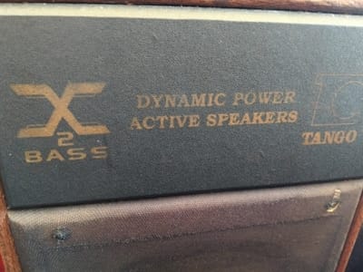 The image shows a portion of a speaker cabinet, specifically the upper front panel. The text on the panel reads "XC Bass", "DYNAMIC POWER ACTIVE SPEAKERS," and "TC TANGO" in gold lettering on a dark grey background. The lower portion of the image shows the speaker grill cloth, which appears worn and slightly damaged, with a small tear. The cabinet appears to be framed in wood.
