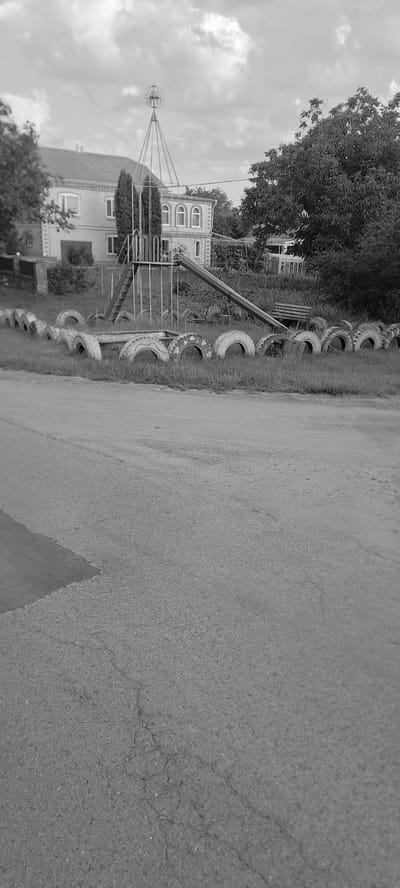 The image depicts a playground area in Trostyanets, Ukraine, with a building in the background. The playground features a metal slide with a ladder, a merry-go-round structure topped with a globe-like ornament, and a bench. The perimeter of the playground is lined with old tires. The scene is set against a backdrop of a two-story building with arched windows and a tree. The foreground shows a cracked asphalt road. The image is in black and white and appears to have been taken during the daytime under an overcast sky.