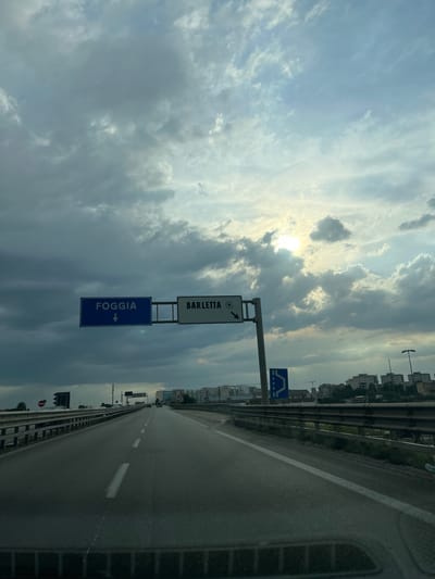 An eye-level shot captures a highway scene under a cloudy sky with the sun partially visible. The camera is positioned inside a vehicle, looking forward along a paved road with visible lane markings. Two overhead road signs are present; the left sign is blue with white text "FOGGIA" and a downward arrow, indicating a direction. The right sign is white with black text "BARLETTA" and an arrow pointing to the right, suggesting the location of Barletta. The road is bordered by a metal guardrail on the right, and on the left, a less visible guardrail. Beyond the guardrail on the right, there are buildings and streetlights. The sky is filled with a mix of dark and light clouds, with the sun's rays breaking through, creating a dramatic effect. In the distance, buildings and trees are visible. The overall impression is one of a slightly overcast day on a highway leading towards a city.