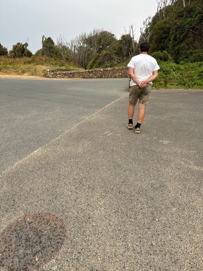 A man wearing a white t-shirt, camouflage shorts, black socks, and athletic shoes stands on an asphalt surface with his back to the camera. He has his hands clasped behind his back. The asphalt is divided by a diagonal line, with one side appearing darker than the other. In the lower left corner of the image, there is a circular, metal manhole cover. Behind the man, a stone wall separates the asphalt from a vegetated hillside with trees and bushes. The sky is overcast. The location is Courtown, Ireland.