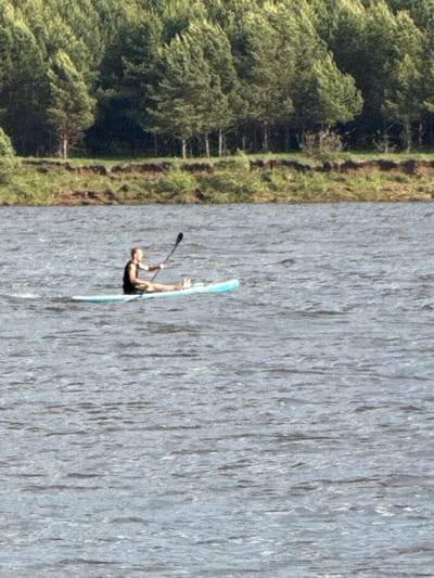 The image shows a person on a paddleboard on a body of water. Lush green trees and vegetation line the bank to the left. The location appears to be Doksha, Russia. The person is using a paddle to move through the water. The water surface is textured, suggesting ripples or slight waves. The image is taken from an aerial perspective and rotated 90 degrees counter-clockwise.