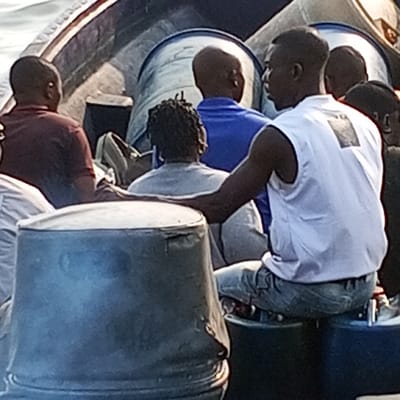 The image depicts at least seven individuals on a boat in Koko, Nigeria. In the foreground, a dark-skinned male wearing a white sleeveless shirt and light-colored jeans sits on a dark blue cylindrical container, his left arm resting on a weathered gray barrel to his left. He is positioned facing right, partially away from the camera. Behind him, other individuals are visible, mostly facing away: one person with dreadlocks in a light gray top, another in a blue short-sleeved shirt, and a person in a dark reddish-brown shirt. Multiple large, cylindrical barrels are present within the boat, including the gray barrel in the foreground, the dark blue barrel used as a seat, and several lighter-colored barrels with blue bands in the background. The boat is on a body of water, visible in the upper left, under daylight conditions.