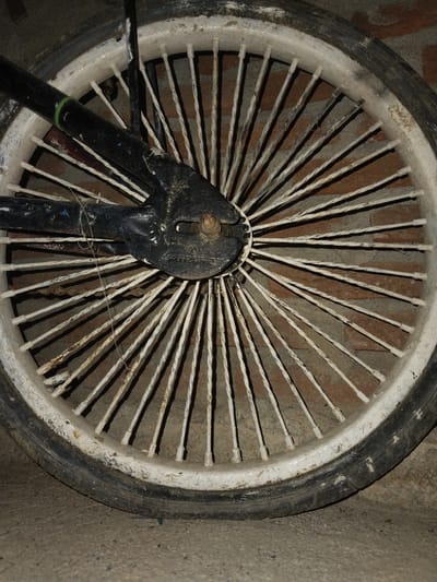 A close-up photograph depicts a bicycle wheel with numerous spokes against a backdrop of bricks and concrete. The tire is black and worn, with a white-painted rim. The spokes are also painted white, showing signs of wear and rust. The center of the wheel is black, attached to the bicycle frame. A stray piece of string or wire is visible near the center. The setting appears to be a building interior. No people or other identifiable objects are present in the image. The photograph does not provide information to confirm or deny its location in Pie, Indonesia.