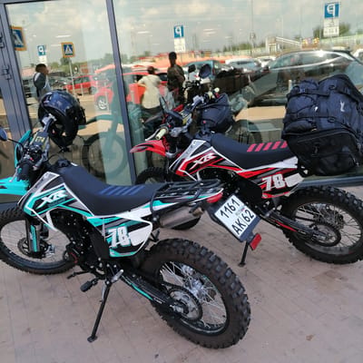 This media file features two dirt bikes parked on a paved walkway in front of a building with large glass windows. The bikes are the primary subjects, with one in the foreground being a turquoise and white model with the number "78" prominently displayed. The second bike, partially visible behind the first, is red and white, also featuring the number "78" on its side. Both bikes appear to be well-maintained and ready for off-road use, with knobby tires and upright handlebars.
In the reflection on the glass windows, a busy outdoor scene is visible, likely a parking lot or plaza. Several cars, including red and silver models, are parked. The reflection also shows people, some of whom appear to be walking or standing. The overall lighting suggests it is daytime, possibly mid-morning or afternoon, with bright sunlight casting reflections on the glass. The weather appears to be fair.
Visible text on the bikes includes brand names like "PROX," "MotoLand," and "BRIDGESTONE," as well as decals with "XR" and "78." One bike has a license plate with the text "1161 RUS AK 62." The environment suggests an urban or suburban setting, with the building and parked vehicles indicating a commercial or public space. The presence of the dirt bikes hints at a hobbyist or recreational context.