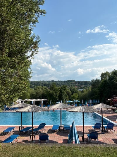 This is a vertically oriented photograph of an outdoor swimming pool area. The pool is filled with clear blue water and is lined with red brick paving. Along the side of the pool are rows of blue lounge chairs and straw-thatched umbrellas, providing shade. The pool is situated at the base of a densely wooded hillside covered in green trees. In the distance, houses are visible nestled among the trees on the hillside, suggesting a village or small town. The sky above is a bright blue with scattered white, fluffy clouds. The lighting indicates it is daytime, likely late morning or early afternoon, with bright sunshine casting shadows from the umbrellas and chairs. There are a few people visible in and around the pool, some swimming and others lounging. The overall atmosphere appears relaxed and leisurely, typical of a summer day at a resort or public pool. Some of the umbrellas have blue and white striped fabric, and one appears to have text on it, though it is too blurry to read. The scene evokes a sense of summer vacation and outdoor recreation.