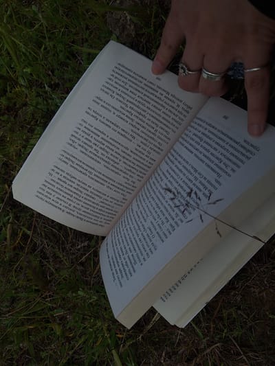 A low-angle, medium shot captures an open book lying on a bed of grass, with a person's hand resting on the right-hand page. The book's pages are filled with text in what appears to be the Russian language. The visible page numbers are 268 and 269. The hand, adorned with multiple rings, holds the book open. The grass is dense and uneven. The lighting is subdued. There is no indication of the location being in the city of Tlik, Armenia.