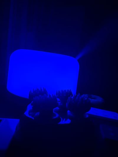 The image, taken in the city of McMasterville, Canada, depicts a dimly lit environment dominated by a vibrant blue light. The primary subject is a rectangular, translucent object with rounded corners, glowing intensely with this blue illumination. In front of this object, and partially obscured by shadows, are several dark, spiky structures that appear to be artificial, possibly decorative elements or part of a display. A faint beam of blue light emanates from the top right of the glowing rectangle, projecting upwards and to the right, suggesting a light source or projection. The overall scene is characterized by high contrast between the bright blue light and the surrounding darkness. No people are visible in the frame.