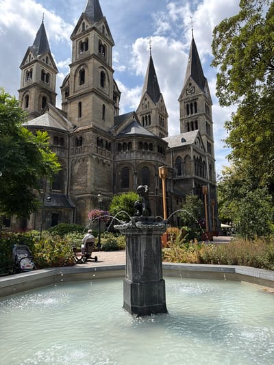 In Roermond, Netherlands, a light-colored fountain with spraying water occupies the foreground. Behind it, a person sits on a mobility scooter in a garden area with lush greenery and blooming flowers. A baby stroller is parked nearby. The main subject is a large, old stone church with multiple spires, Romanesque architectural features, and a clock on one of its towers. Trees frame the right side of the image, and a cloudy sky is visible overhead.