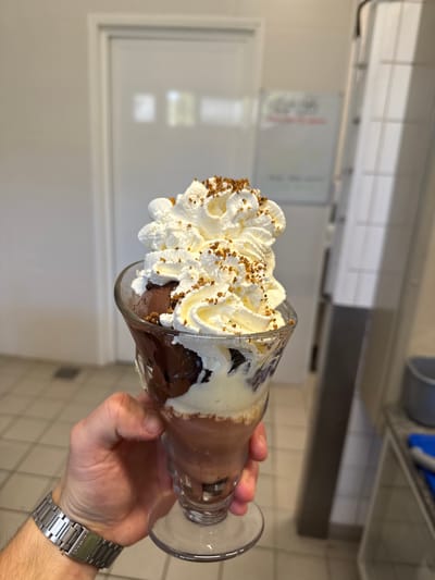A person's hand, wearing a silver metal watch, holds a clear glass goblet filled with a chocolate and vanilla ice cream sundae. The sundae is topped with a generous amount of whipped cream and sprinkled with small brown crumbles. The background appears to be an indoor setting with tiled floors and a partially visible door and metallic shelving. No specific location in Rivesaltes, France, can be determined from the image.