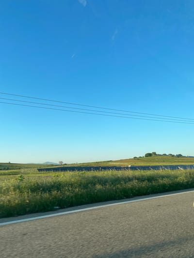 The image shows a road and adjacent field next to a solar panel farm on a sunny day in Myrodato, Greece. The sky is clear and blue. Power lines run parallel to the road.
