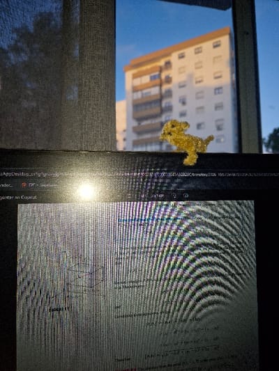 The image displays an indoor scene overlooking an exterior view in Carnaxide, Portugal. In the foreground, the upper section of a dark laptop is visible. Its screen exhibits a pattern of vertical lines and reflections, displaying what appears to be academic material with mathematical equations, text, and a geometric diagram. A small, fuzzy, yellow, dog-shaped decorative item is perched on the upper bezel of the laptop. Behind the laptop, a window reveals an outdoor environment. A dark, textured curtain or blind obscures the left portion of the window frame. Through the unobstructed window area, a multi-story residential building with a light-colored facade and a prominent brown-orange upper floor or roof section is visible under a clear blue sky. The building is distinctly illuminated by direct sunlight. Sparse dark foliage is present on the periphery of the outdoor view. No individuals are depicted.