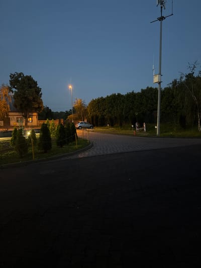 The image depicts an outdoor scene at dusk in Wąwelnica, Poland, likely a residential or public area, possibly a parking lot or roundabout. The sky is a deep twilight blue, with a single streetlamp casting a warm orange glow. In the background, a silver car is parked near a building with lit windows. A tall utility pole with antennas and a white box is prominent on the right side of the frame. Several small, cone-shaped evergreen trees are arranged in a line, bordered by a low fence. The ground is paved with interlocking bricks, and the foreground is dark asphalt. Vegetation, including trees and shrubs, surrounds the paved area. There are no people visible in the image. The overall impression is one of quietude and transition from day to night.
