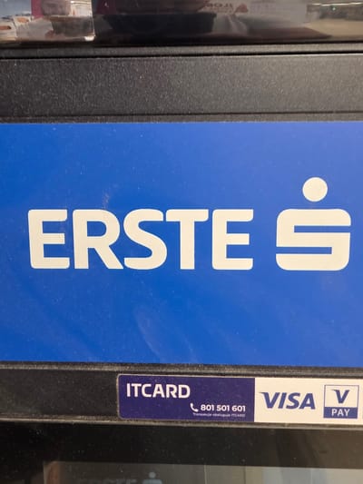 The image shows a close-up of an ATM machine. The ATM displays the logo for "ERSTE" bank, which features white lettering on a bright blue background. Below the "ERSTE" logo, there is a strip with several labels. On the left of this strip, the word "ITCARD" is visible in white text on a dark blue background, along with a phone number "801 501 601" and a line of text in Polish. To the right of "ITCARD", the "VISA" logo is present in blue lettering on a white background, followed by the "V PAY" logo. The ATM is located in Brzeg, Poland. No people are visible in the image, and the environment appears to be indoors, likely a public space such as a bank branch or shopping center.