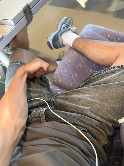 The image shows a close-up view of a person's legs and arm, seated on a purple and white patterned surface, likely a seat on public transportation. The person is wearing denim shorts, a grey t-shirt, white socks, and grey athletic shoes. A white cable, possibly from headphones or a charger, is visible across the person's lap. The background appears to be the floor of a vehicle, with shadows suggesting movement or an uneven surface. The image does not provide any specific information or visual cues to indicate the location within the city of Rivesaltes, France.