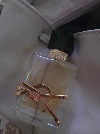 A clear glass perfume bottle with a dark, square-shaped cap and a gold-textured band is depicted. A prominent gold-colored, stylized logo, identifiable as "YSL," is affixed to the front of the bottle, which contains a light amber liquid. The bottle is positioned diagonally, partially submerged within an open light-colored, textured bag, likely made of synthetic leather. The bag features visible stitching along an edge and a silver-toned metal eyelet. A portion of a patterned surface, possibly textile, is visible beneath the bag. No individuals or active events are present in the image, which is located in Staliyska mahala, Bulgaria.