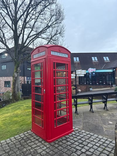 A bright red, traditional British K6 telephone kiosk stands prominently on a cobblestone surface in a public area of Bordon, Whitehill, East Hampshire, United Kingdom. The kiosk features "TELEPHONE" signage and royal crown emblems above its glass-paneled door, marked with a "PULL" handle. To the left, a grassy slope rises towards a large, bare deciduous tree, behind which a dark-clad building with multiple windows is visible, partially obscuring a sign reading "PRG SHOP". To the right, a paved walkway leads to two empty black park benches with ornate supports. Further right and behind the benches, a multi-directional wooden signpost stands before a building housing "Oties" cafe, identified by a large glass facade and signage for "WAFFLES • COFFEE." The building's roof features several dormer windows. The sky is overcast, indicating a dull daylight condition. No individuals are present in the scene.