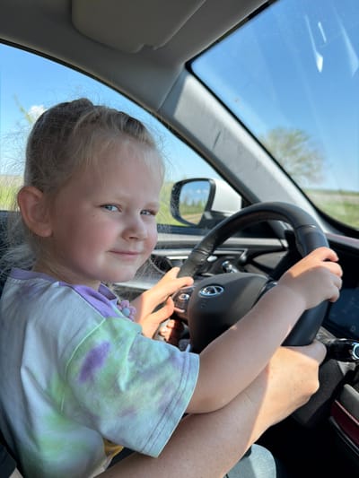 A young girl is seated in the driver's seat of a car, holding the steering wheel. An adult's hand is also on the steering wheel. The girl is fair-skinned with blonde hair. She is wearing a tie-dye shirt. The car is in motion as scenery is visible through the windshield. The location is not specified or identifiable in the image.