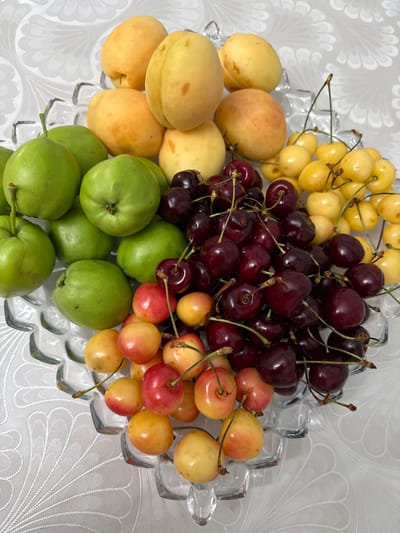 A clear glass bowl filled with a variety of fresh fruits sits on a white tablecloth with a delicate, embossed floral pattern. The bowl is overflowing with a colorful assortment of cherries, apricots, and green apples. 

The cherries are a mix of deep red and lighter yellow varieties, some still attached to their stems. The apricots are a soft orange-yellow hue, plump and smooth. The green apples are small and round, with a vibrant green skin. The fruits are arranged in a natural, abundant display, suggesting a recent harvest or preparation for a meal.

The lighting is bright and even, indicating an indoor setting with good illumination, possibly during the daytime. There are no people visible in the image. The overall impression is one of freshness, health, and abundance. The setting appears to be a home, given the tablecloth and the domestic presentation of the fruit. The location context provided is Artik, Armenia.