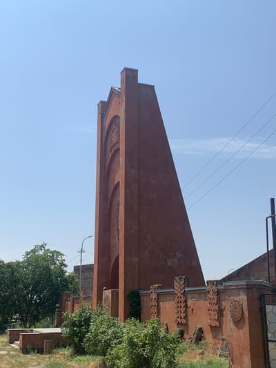 The image shows a monument in the village of Karbi, Armenia. The monument is made of red stone and has a tall, triangular shape. It is decorated with arched carvings. A low wall with intricate stone carvings surrounds the monument. Trees and bushes are visible in the foreground and background. The sky is clear and blue. There are no people or animals in the image. The scene is set outdoors on a sunny day.