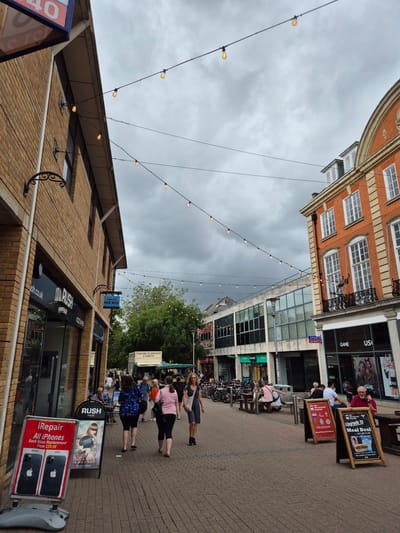 This is a street-level, eye-level, medium shot capturing a pedestrian area in Cambridge, United Kingdom, likely a shopping street, under an overcast sky. A prominent row of shops lines the right side of the street, featuring architectural styles ranging from traditional brick to modern glass facades. The buildings are adorned with a string of decorative lights, suggesting an evening or festive atmosphere, though the daylight indicates it is daytime.
On the left, a brick building with windows and awnings houses businesses such as "RUSH HAIR" and "iRepair," indicated by signage and promotional boards. The "iRepair" board displays an advertisement for iPhone back glass replacement and features images of iPhones.
The street itself is paved with brickwork, and a variety of people are walking or sitting in outdoor seating areas associated with the establishments. There are benches and tables outside some of the shops. On the right side, a prominent orange brick building with arched windows and decorative balconies stands out. Shops on this side include "GAME" and "US."
In the middle ground, a "FRESH FLOWERS & FRUIT" stall is visible, and behind it, a dense cluster of trees forms a green backdrop. Bicycles are parked in racks in the distance.
Several individuals are the subjects of the scene. Two women in dark leggings and tops are walking away from the camera, one in a blue patterned top and the other in a pink top carrying a black bag. Another woman in a blue dress walks further away. A man in a red shirt is seated at a table on the right, holding a phone to his ear and looking towards the camera. Other people are visible in the background, either walking, sitting, or browsing. The overall impression is a moderately busy urban environment.