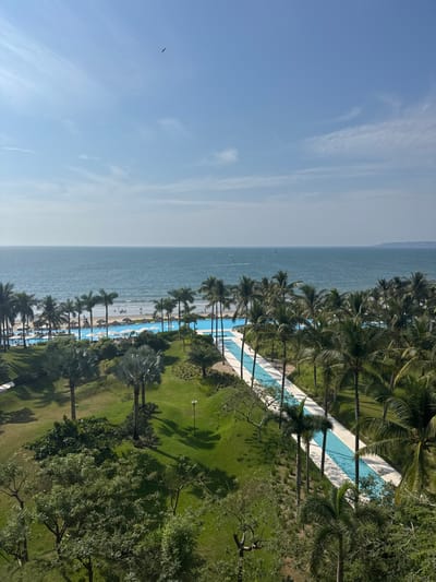 A coastal vista showcases an azure ocean meeting a sandy beach fringed with swaying palm trees. Lush green lawns and a meandering, bright blue swimming pool form part of a resort landscape. The clear, light blue sky complements the scene. The composition balances natural elements with manicured spaces.