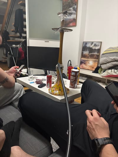 The image depicts an indoor scene within a dwelling in Krems an der Donau, Austria. At least three individuals are partially visible, seated on a grey upholstered sofa. On the left, a person in a light grey long-sleeved top and grey sweatpants has a tattoo reading "hope" on their right forearm and is holding a dark smartphone with both hands. Their feet, clad in dark socks, are visible. In the immediate foreground, an individual is seen wearing black sweatpants. To the right, another person wears a dark digital watch on their left wrist and holds a dark smartphone vertically.
A tall hookah, featuring a yellow and white speckled glass base, a wooden stem, and a clear glass bowl with shisha tobacco and foil/charcoal, stands centrally on a white rectangular coffee table. The hookah's dark hose extends towards the foreground. Also on the table are a clear drinking glass containing a clear liquid, a blue cigarette lighter, two red-and-white cigarette packs, a black eyeglasses case, a red Coca-Cola can, and a brown carton of "RÖMÜ Kakao Milch" (cocoa milk), alongside a clear glass ashtray with residual ash.
In the background, a large dark monitor rests on a white console. To the left, a wooden open-tread staircase with clothing hanging on a drying rack is visible. Two framed artworks, depicting a waterfall and a landscape, are mounted on the white wall. A stack of folded blankets or textiles is located on a shelf to the right.