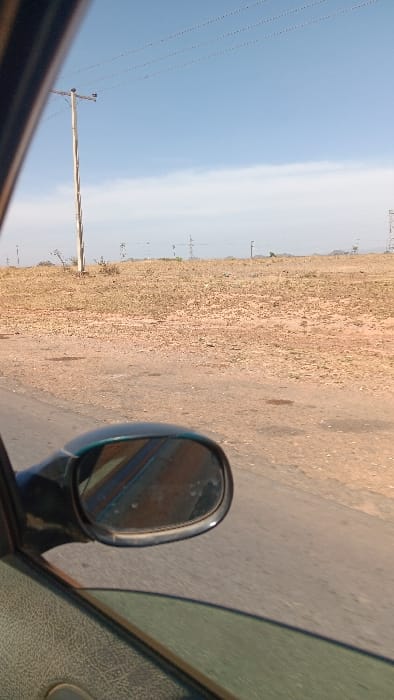 The image displays a roadside scene in Barakin Columbai, Nigeria, captured from inside a moving vehicle. The foreground on the left shows a portion of a dark, textured car door panel and the lower edge of a partially open window with a green tint. A dark-colored side mirror of the vehicle is prominent in the lower-middle, reflecting a blurry, reddish-brown image. Adjacent to the vehicle is a grey paved road. Beyond the road, a wide, dry expanse of reddish-brown and light brown earth dominates the mid-ground, characterized by sparse, dried vegetation. To the left, a tall, light-colored wooden utility pole stands, supporting multiple overhead power lines that extend horizontally across the bright blue sky. Further in the distance, towards the right, several metallic electrical transmission towers are visible against the horizon. The sky is predominantly clear blue, with a faint horizontal band of white cloud across the middle. Distant hills are faintly discernible on the horizon. No people are visible.