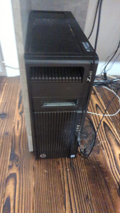 The image shows a black HP Z440 workstation computer tower on a wooden floor. The computer tower is oriented vertically, with its front facing the viewer. The top of the tower has a textured surface. The front of the tower has several horizontal ventilation slats, an optical drive bay with a metallic front, and the HP logo. The side of the tower has the "Z440" model number printed vertically. Several black cables and one white cable are visible, connecting to the back or side of the computer and running across the floor. The wooden floor has a pronounced grain pattern and appears to be made of planks. The background is out of focus but suggests a wall or partition. There is no visible indication of the city of Zadebri, Ukraine, or any human presence.