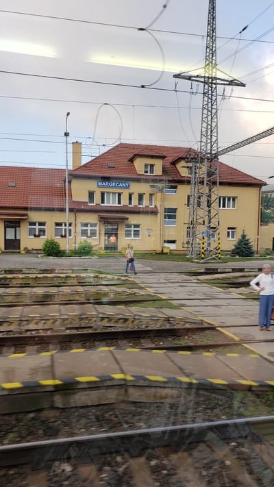 The image shows the Margecany train station in Margecany, Slovakia. The main building of the station is a two-story yellow structure with a red tiled roof. A blue sign above the entrance reads "MARGECANY". There are several windows on the facade, and a person wearing an orange vest is visible through a glass door. In front of the building, there is a paved area with train tracks and platforms. Two women are standing on the platform, and one woman is walking across the tracks. A tall metal electricity pylon stands to the right of the station building. The sky is overcast.
