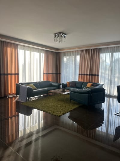 A well-lit, modern living room in Brca, Montenegro features a large, dark green leather sectional sofa arranged in an L-shape around a glass-topped coffee table. A vibrant green shag rug sits in the center of the seating area. The room is illuminated by natural light streaming through large windows covered by sheer white curtains and accented with floor-to-ceiling curtains in shades of peach and brown. A contemporary silver chandelier hangs from the ceiling. The room's floor is made of highly polished, dark marble tiles that reflect the light and the furnishings. On the sofa, several throw pillows in various colors, including yellow, orange, and brown, are visible, along with a striped throw blanket draped over one section of the sofa. The environment appears clean and organized, with no people present.