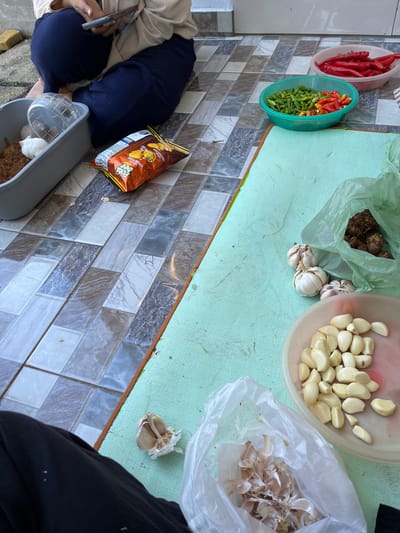 The image depicts an outdoor scene on a tiled floor, likely in Jambearum, Indonesia, where someone is preparing ingredients. A person, partially visible and wearing dark blue pants and a cream-colored top, is seated and looking at a smartphone. In the foreground, several food items are laid out on a light green mat. These include bowls of red and green chilies, peeled garlic cloves in a pink bowl, and unpeeled garlic bulbs. A plastic bag contains what appear to be ginger or turmeric roots. A bag of potato chips rests on the tiled floor near a gray container holding a small white rodent and some dried material. The overall impression is of a casual, domestic setting where food preparation is underway.