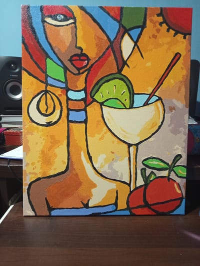 This is a close-up, eye-level shot of an abstract acrylic painting on canvas, which is leaning against a dark wooden surface. The painting depicts a stylized female figure with bold black outlines and vibrant colors, primarily oranges, yellows, reds, blues, and greens. To the left of the painting, a black studio monitor speaker is visible. The background of the painting features abstract shapes and patterns. A margarita glass with a lime slice and a straw is prominently featured to the right of the figure. Below the glass, two red fruits, possibly apples or cherries, are painted. The overall impression is a lively and colorful composition with a modern, cubist influence. The painting is likely displayed in a home or studio setting, given the presence of the speaker and the dark wooden surface which could be a desk or table. There is no indication of the specific location in the city of Чэрвеньскі сельскі Савет, Belarus.