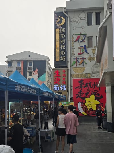 This is an eye-level, outdoor, medium shot of a street scene in Gongshu District, China, under an overcast sky. The foreground is dominated by blue market tents, likely for food vendors, with white lettering that appears to be in Chinese characters. A woman in a black shirt is standing behind a stall, partially obscured by a white object and blue cooler. To her left, a person's head wearing a white cap is visible. In the midground, several people are walking down the street. A young couple, a girl in a white top and black skirt, and a boy in a pink t-shirt and navy shorts, are walking away from the camera. Behind them, a young boy in a striped shirt walks towards the right side of the frame. Further back, a security guard in a blue uniform is standing near a trash can. The background features a multi-story building with a large mural of a woman's face with striking orange and teal accents. Above this mural, a tall vertical sign with Chinese characters in gold and black is prominent, featuring a crescent moon design at the top. To the right of this sign, another building displays colorful Chinese characters that appear to be advertisements or store names. A large red sign with black Chinese calligraphy and a cartoonish yellow star with a fish is also visible on this building. The street pavement is a light grey tiled surface.