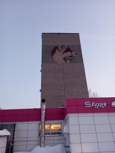 The image depicts an urban winter scene in Яромаска, Russia, featuring two primary structures. In the background, a tall, multi-story rectangular concrete building, likely an apartment block, stands against a light blue sky. Its upper facade displays a large, circular abstract mosaic art piece composed of geometric shapes in muted red, grey, and brown tones. In the foreground, a lower, modern commercial building features a prominent pink/magenta fascia and white tiled walls. A white sign on the pink fascia reads "M Кухня" in Cyrillic letters, with a stylized "M" logo preceding the word. A tall, corrugated metal chimney extends vertically from the roof of the foreground building, passing a window with illuminated interior lights. Snow accumulates on the ground and on the roof surfaces of the foreground structure. No individuals are visible. The overall environment suggests cold weather conditions.