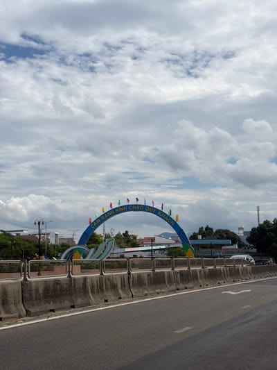 The image depicts an outdoor street scene in Cam Ranh Ward, Vietnam, dominated by a large welcome archway under a partly cloudy sky. The perspective is angled, showing a road on the right side of the frame and the archway and surrounding landscape on the left.
An asphalt road runs along the right, featuring a solid white line at its edge and a painted white arrow indicating the direction of traffic. A robust concrete barrier, topped with metal railings, separates the roadway from the adjacent area.
To the left of the barrier stands a prominent blue archway, possibly a ceremonial entrance or welcome gate to Cam Ranh. This archway is circular or semi-circular and adorned with a string of small, colorful flags. Clearly visible text on the archway reads "CAM RANH CHÀO QUÝ KHÁCH," which translates to "Cam Ranh Welcome Guests." Behind and around the archway, various buildings are visible, including some with industrial characteristics and others with residential roofs, interspersed with lush green trees. A tall antenna or communication tower can be seen rising in the distance.
The sky, occupying a large portion of the upper and left side of the image, is filled with a mix of white and grey cumulus clouds, with occasional glimpses of blue sky between them. The overall lighting suggests it is daytime, likely late morning or early afternoon. There are no people visible, and no specific activities are taking place, presenting a static view of the local infrastructure and landscape.
