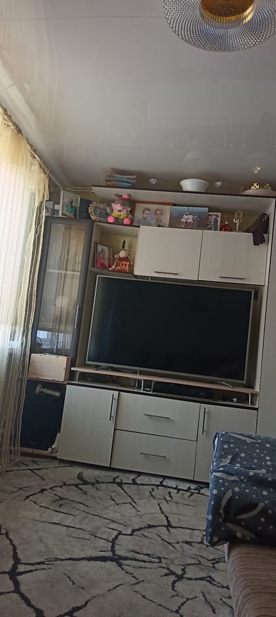 The image depicts an interior living room scene. Dominating the center is a large flat-screen television mounted on a media console. The console has several drawers and cabinets. Above the television, a shelf displays various decorative items, including framed photographs, stuffed animals, and trophies. To the left of the media console is a tall glass-fronted cabinet, also containing displayed items. A beige curtain hangs to the far left of the image, partially obscuring a window. The floor is covered with a patterned rug. The ceiling features a light fixture. The overall impression is of a domestic living space, likely a living room.