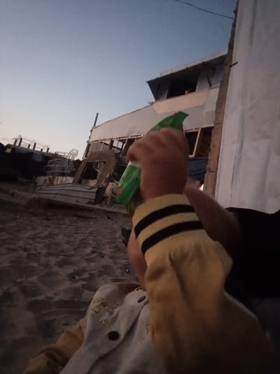 The image depicts an outdoor scene at dusk or dawn in Al-Mawasi, Palestinian Territories. In the mid-right foreground, a person's arm, covered by a yellow sleeve with two black horizontal stripes, is holding a green plastic object, possibly a spray bottle, which displays faint red markings. Partially visible below this arm is the upper torso of a child wearing a light-colored garment with dark buttons and embroidered details. The ground consists of textured sand. In the background, under a sky transitioning from dark blue to a lighter hue near the horizon, several damaged structures are present. To the left, corroded, skeletal metal structures, suggestive of industrial wreckage or boat remnants, are visible. In the center-right, a large, light-colored building, exhibiting significant structural damage and featuring corrugated metal components, contains multiple empty window openings. A vertical white tarp or fabric stands to the far right, partially obscuring this building. The perspective is low-angle, looking upwards.