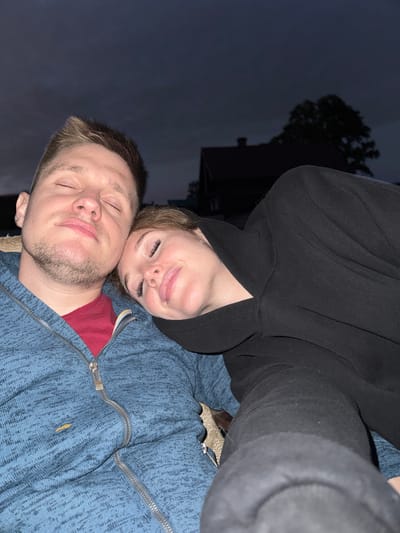 In Maksimovo, Russia, at dusk, a man and a woman are pictured resting their heads together, eyes closed, with soft smiles on their faces. The man, wearing a blue zippered hoodie over a red shirt, has short brown hair and stubble. The woman, nestled against him, is wearing a black hooded sweatshirt and has light brown hair. The background is dimly lit, showing a silhouette of a house and a tree against a cloudy sky. The overall atmosphere is one of peaceful intimacy and comfort.